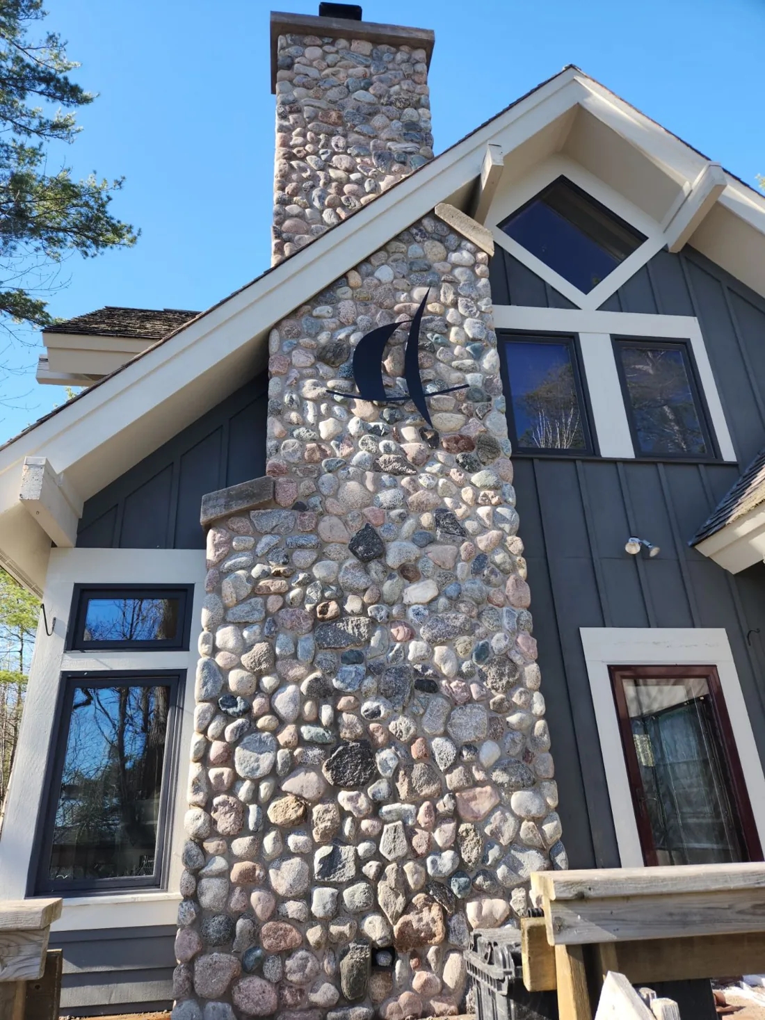 Tall rounded-stone chimney on a gray house with a sailboat ornament on its face