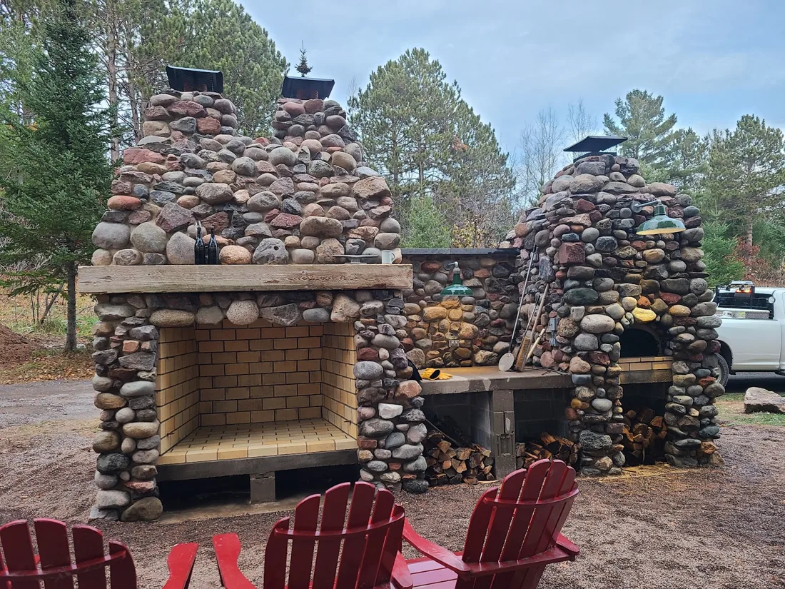 Two rounded-stone outdoor fireplaces with red chairs and trees around them